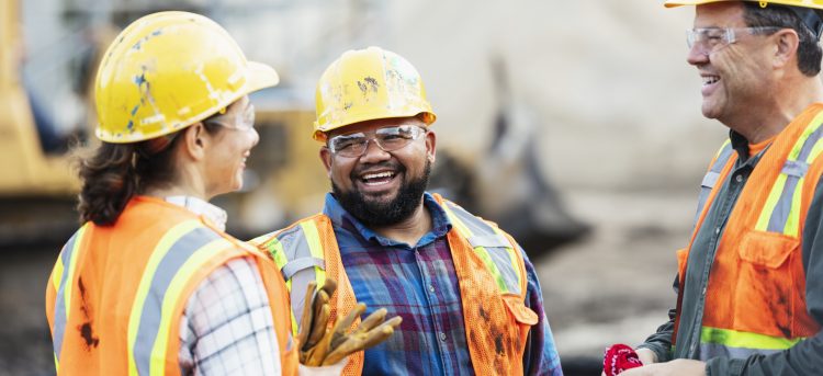 A group of three multi-ethnic workers at a construction site wearing hard hats, safety glasses and reflective clothing, smiling and conversing. The main focus is on the mixed race African-American and Pacific Islander man in the middle. The other two construction workers, including the woman, are Hispanic.